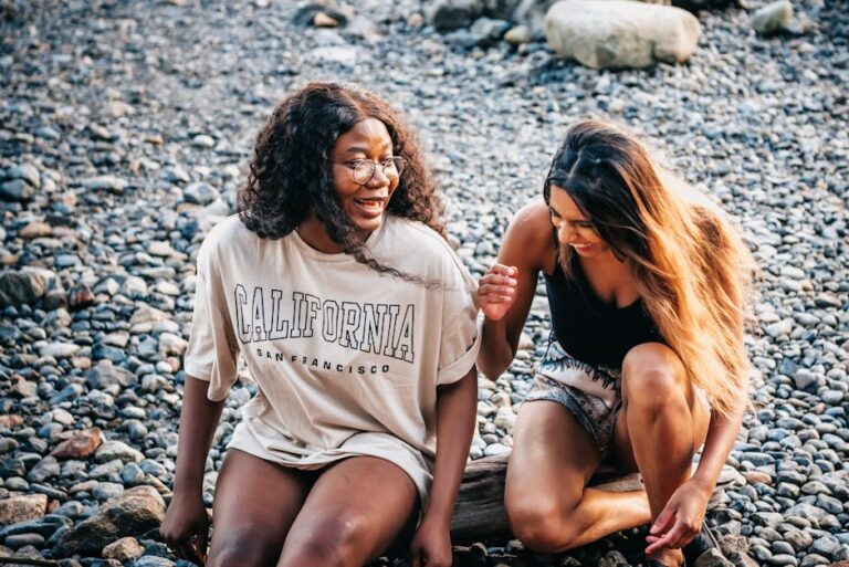 Two women enjoying a joyful moment sitting on a rocky beach, laughing together.
