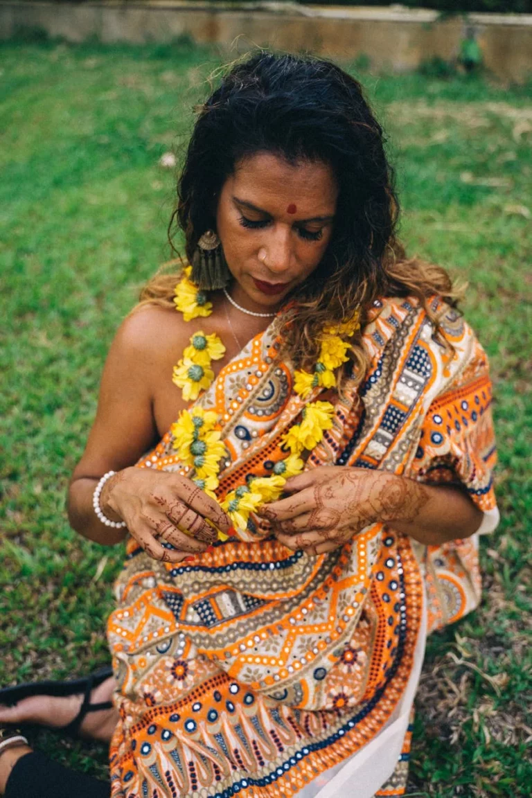 South Asian woman wearing a saree and floral garland, sitting on grass outdoors.