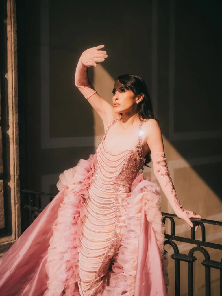 Fashion model poses in a luxurious pink gown with dramatic lighting indoors.