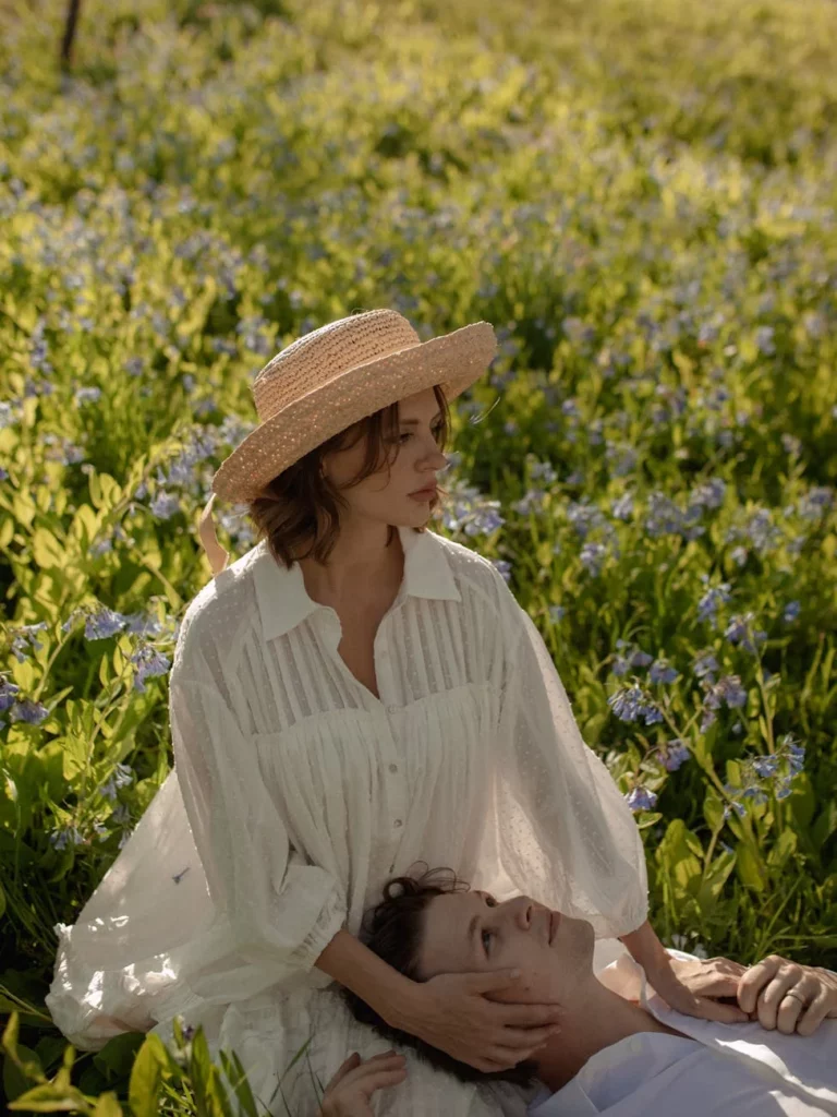 A woman in a straw hat and white dress sits with her partner in a blossoming meadow.