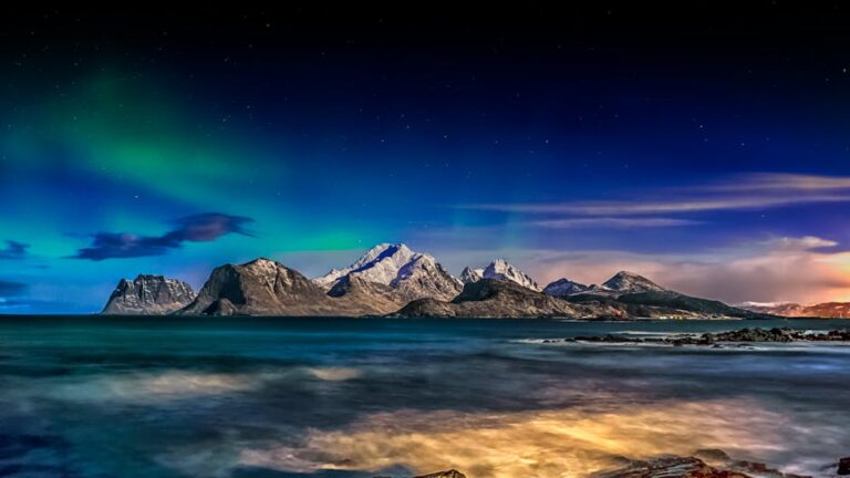 Breathtaking aurora over snow-capped mountains and ocean in Napp, Norway during twilight.